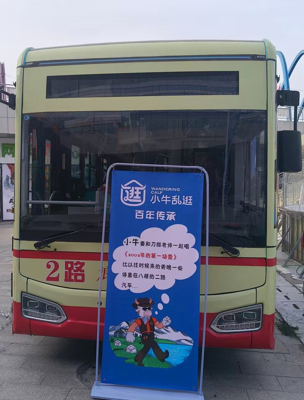 新疆乌鲁木齐，一辆 2 路公交车停在站台，车前立着“小牛乱逛”蓝色立牌，牌上写着改编自《2002 年的第一场雪》的几句歌词