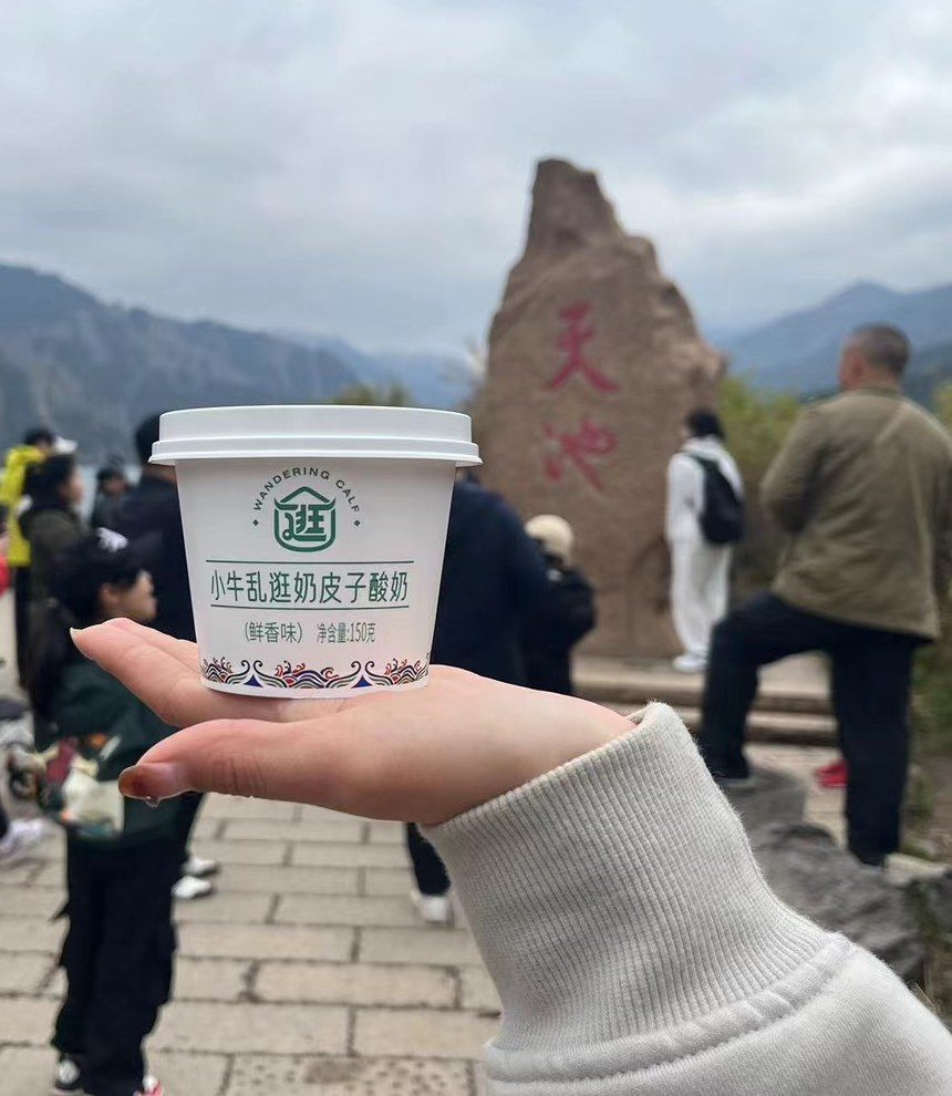 天山天池湖畔，一只手举起小牛乱逛奶皮子酸奶，背后是刻着“天池”二字的题词石。