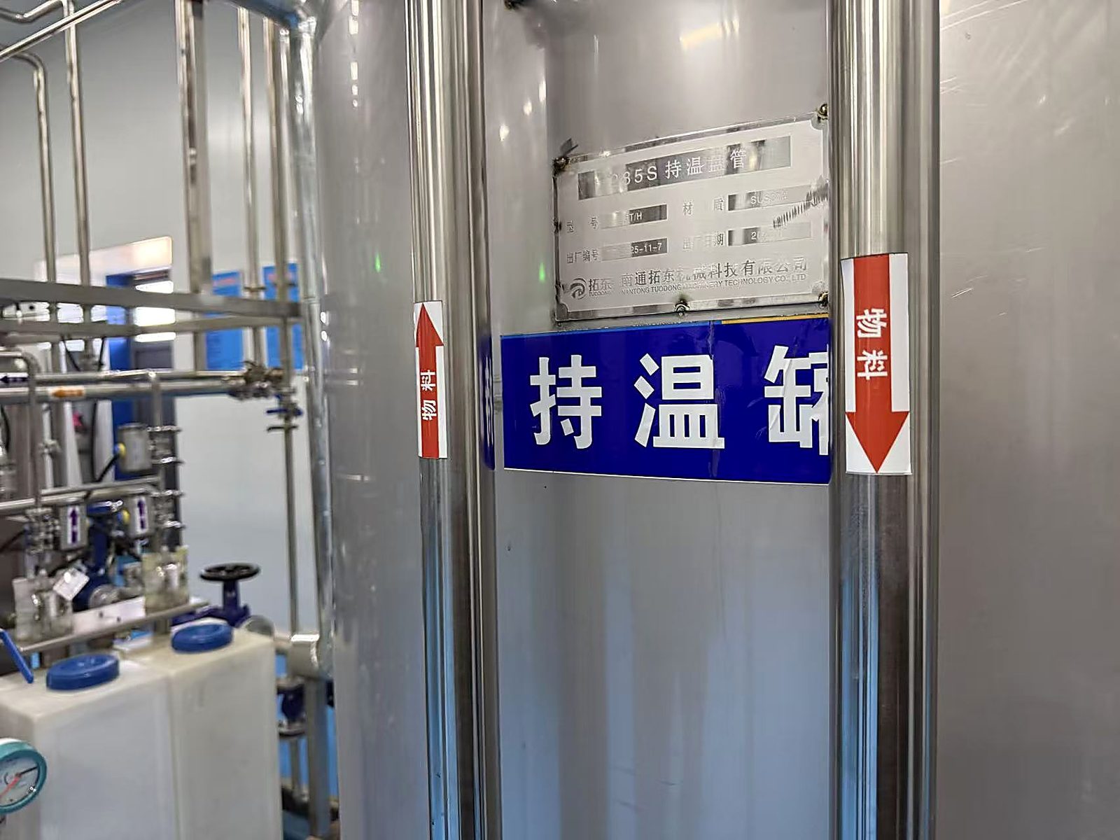 车间内的持温罐与管道。罐体标牌上写着"持温缶"，两侧红箭头标着物料流向；罐体上方的铭牌记录着这台设备的型号与制造企业。拍摄于 2026 年 9 月 26 日。